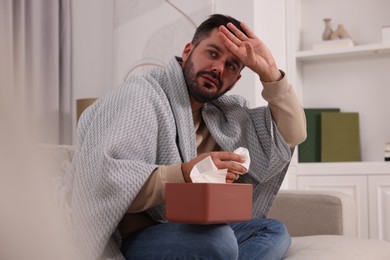 Photo of Sick man wrapped in blanket with tissues on sofa at home. Cold symptoms