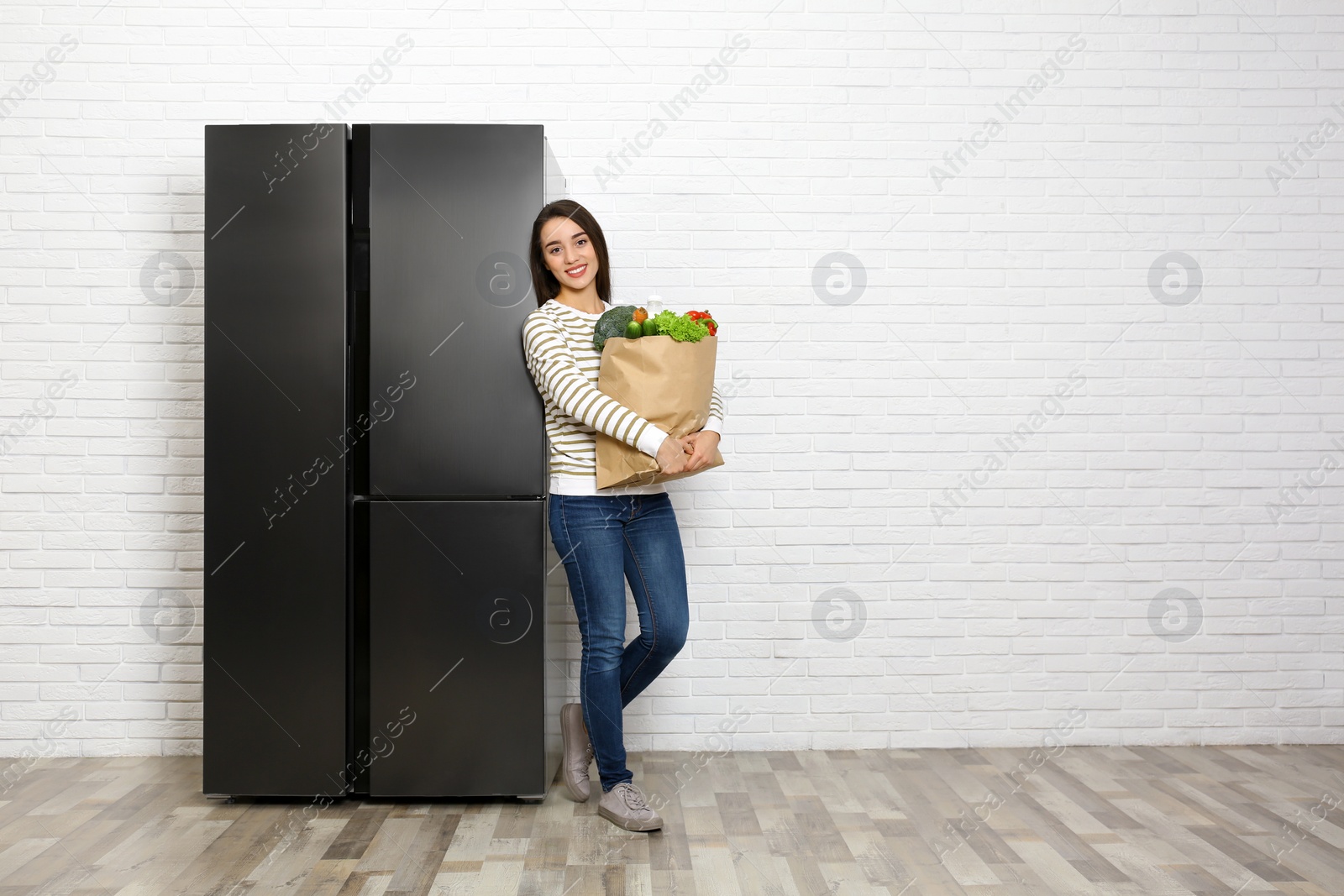 Photo of Young woman with paper bag full of products near refrigerator indoors, space for text