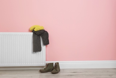 Heating radiator with knitted cap, scarf and shoes near color wall. Space for text