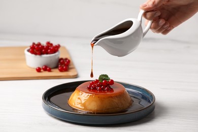 Woman pouring caramel onto delicious pudding with red currants and mint on white wooden table, closeup