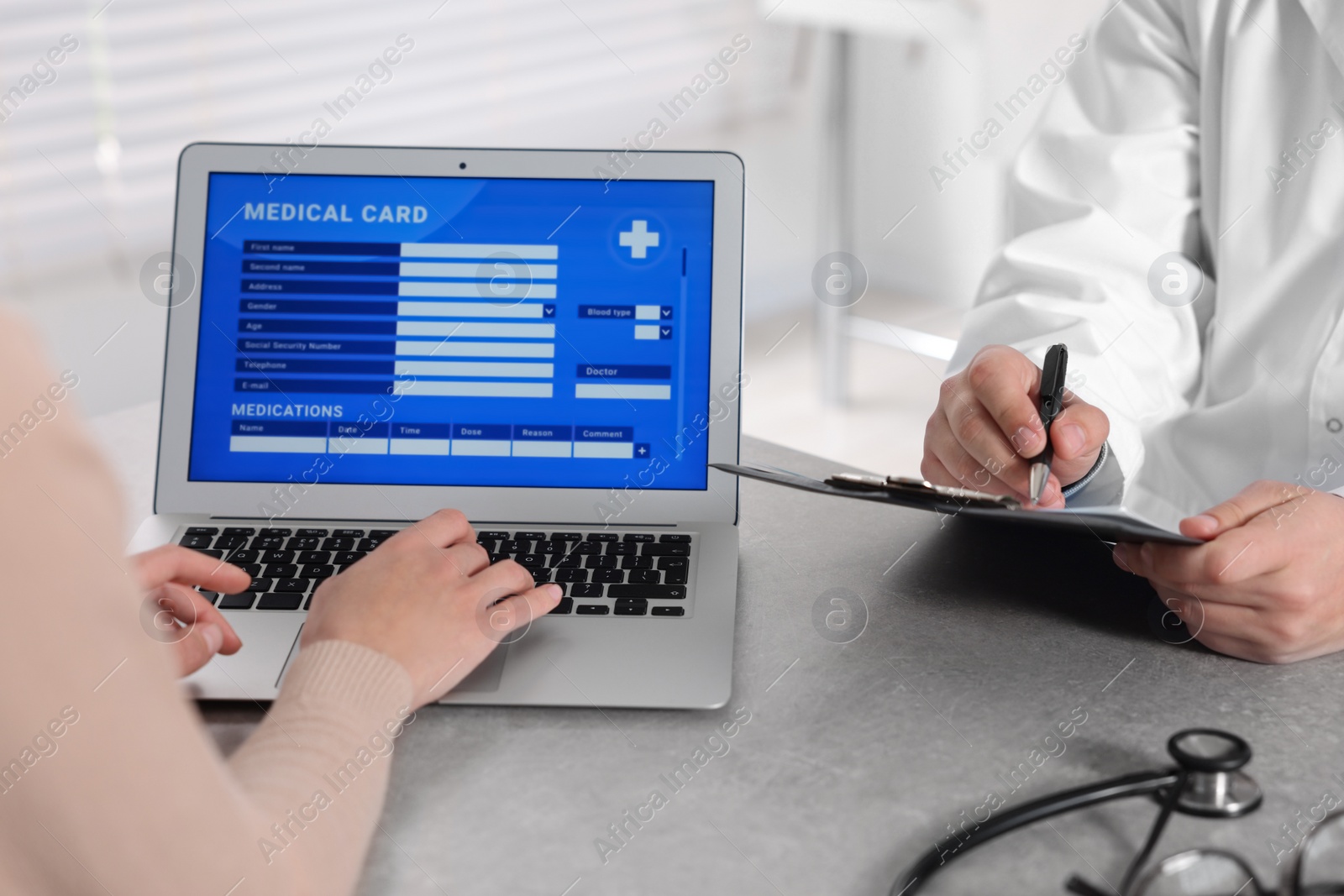 Photo of Doctor and patient using laptop to fill out medical card at table in clinic, closeup