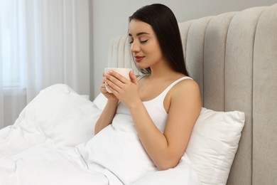 Beautiful young woman with cup of drink in bed at home