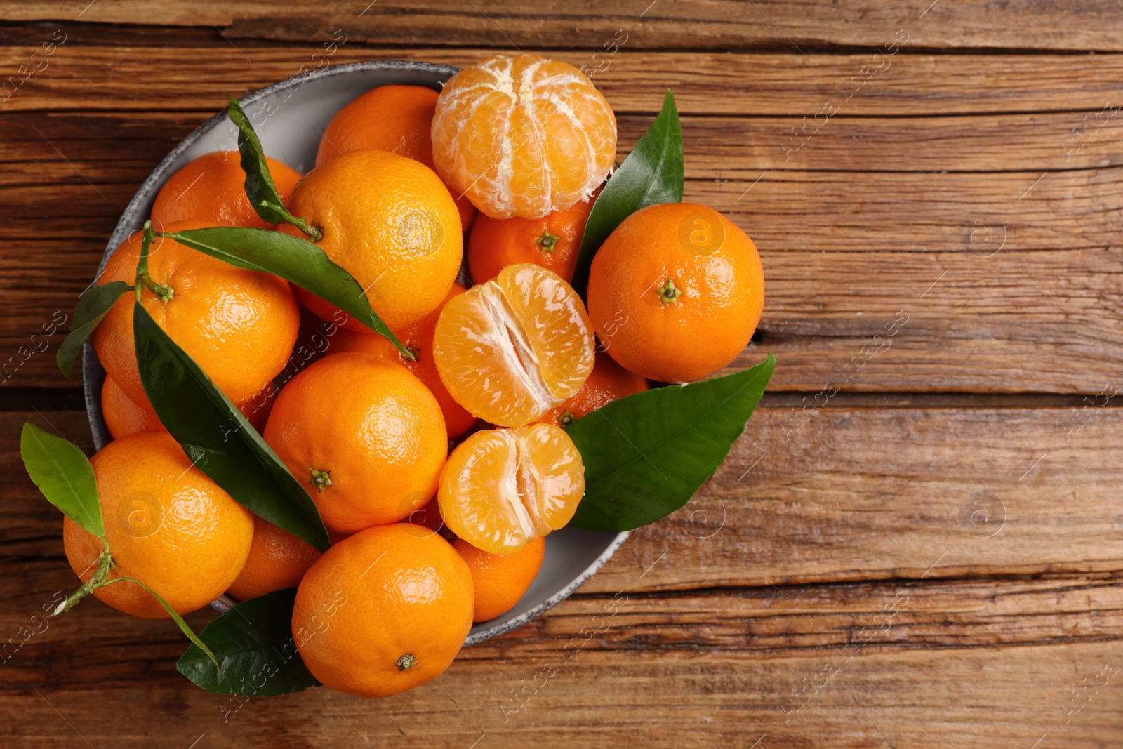 Photo of Fresh tangerines with green leaves in bowl on wooden table, top view. Space for text