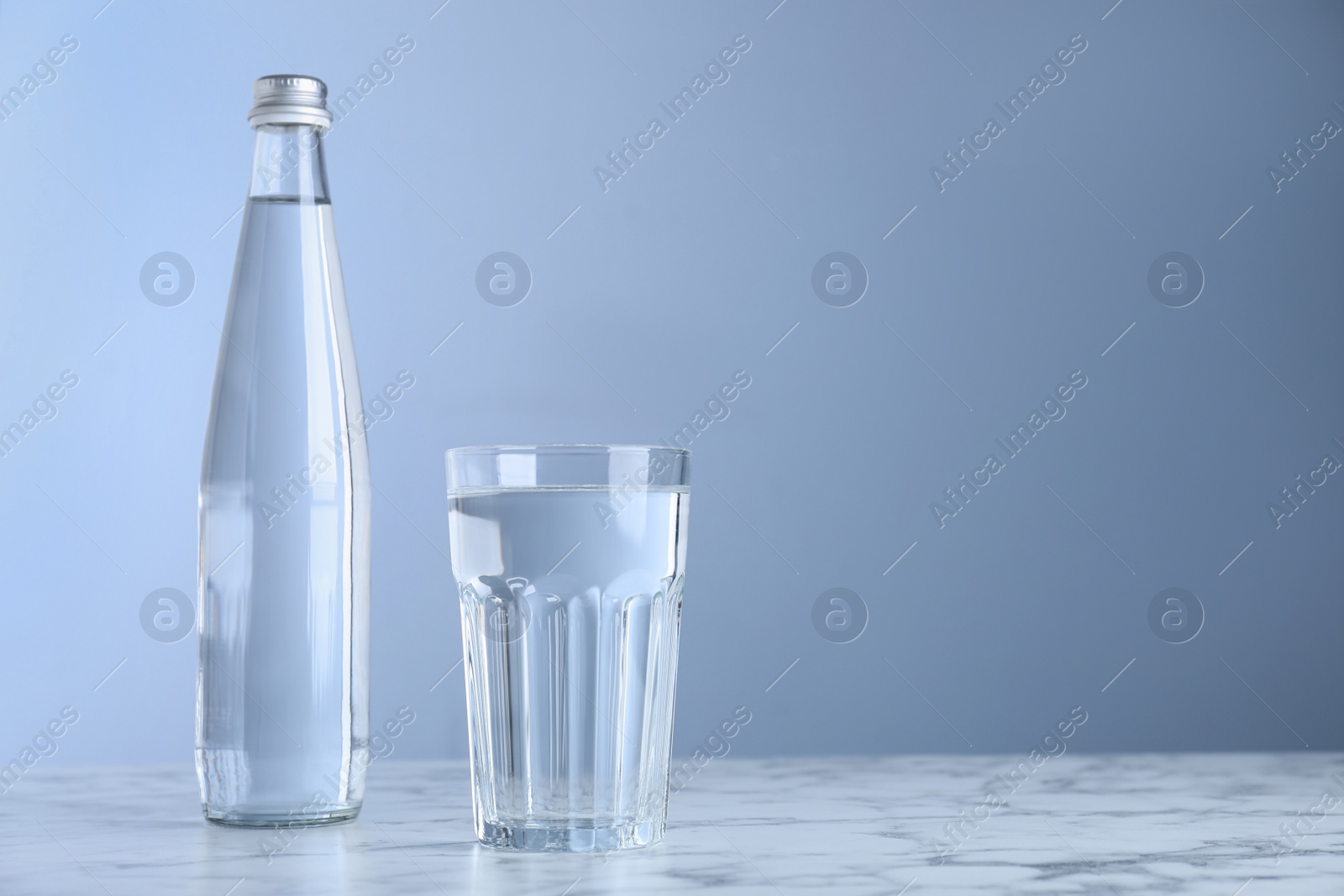 Photo of Glass and bottle with water on white marble table against blue background, space for text