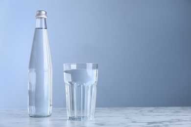 Photo of Glass and bottle with water on white marble table against blue background, space for text
