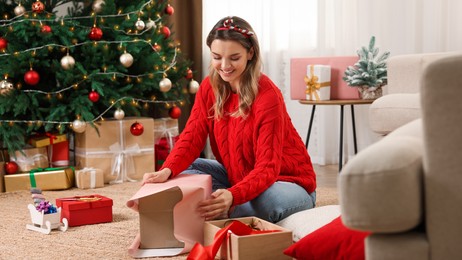 Photo of Beautiful young woman wrapping gift near Christmas tree at home