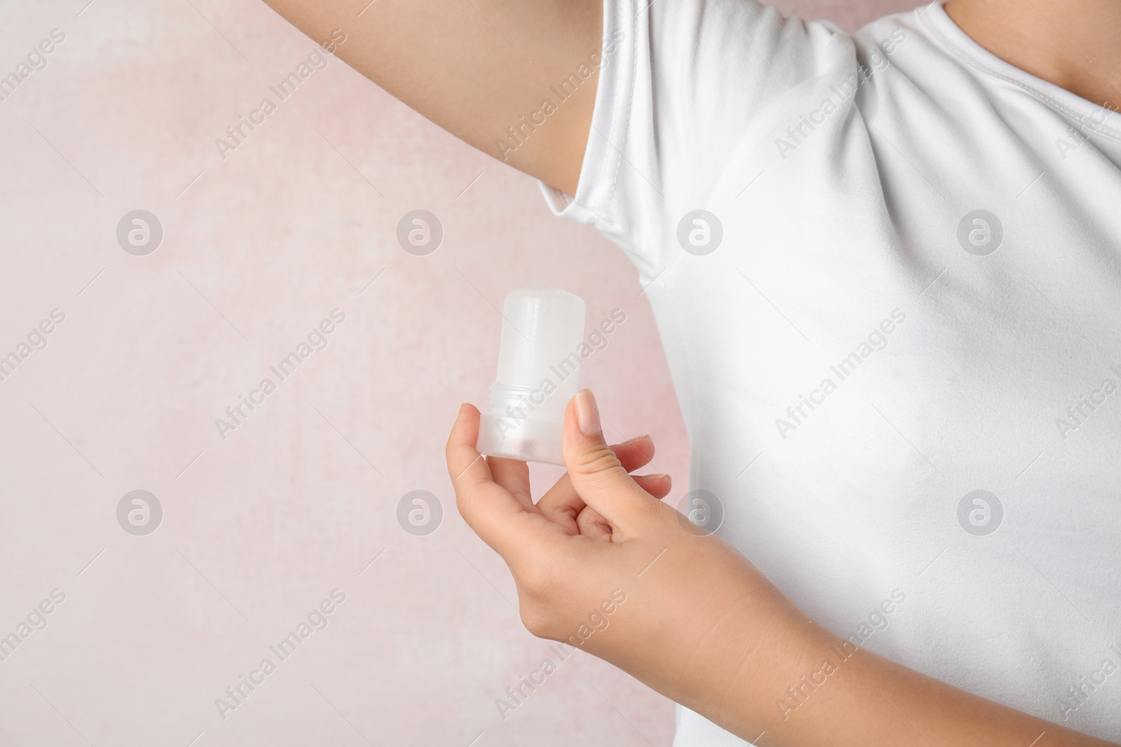 Photo of Young woman holding natural crystal alum deodorant near armpit on light pink background, closeup