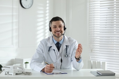Photo of Doctor with headset consulting patient over phone at desk in clinic. Health service hotline