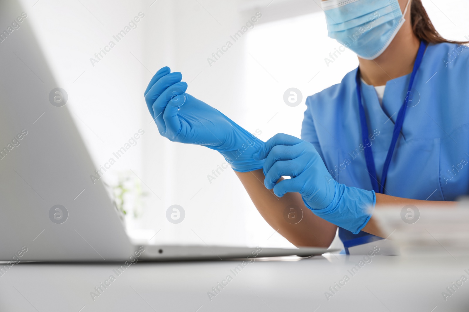 Photo of Doctor in protective mask putting on medical gloves at table in office, closeup