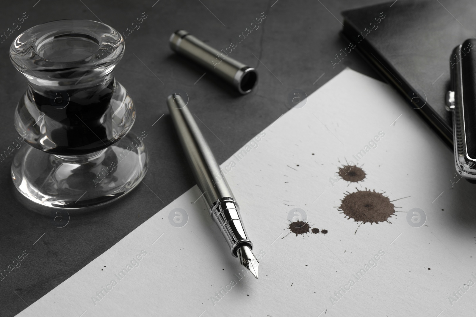 Photo of Stylish fountain pen, paper with blots of ink and inkwell on grey textured table