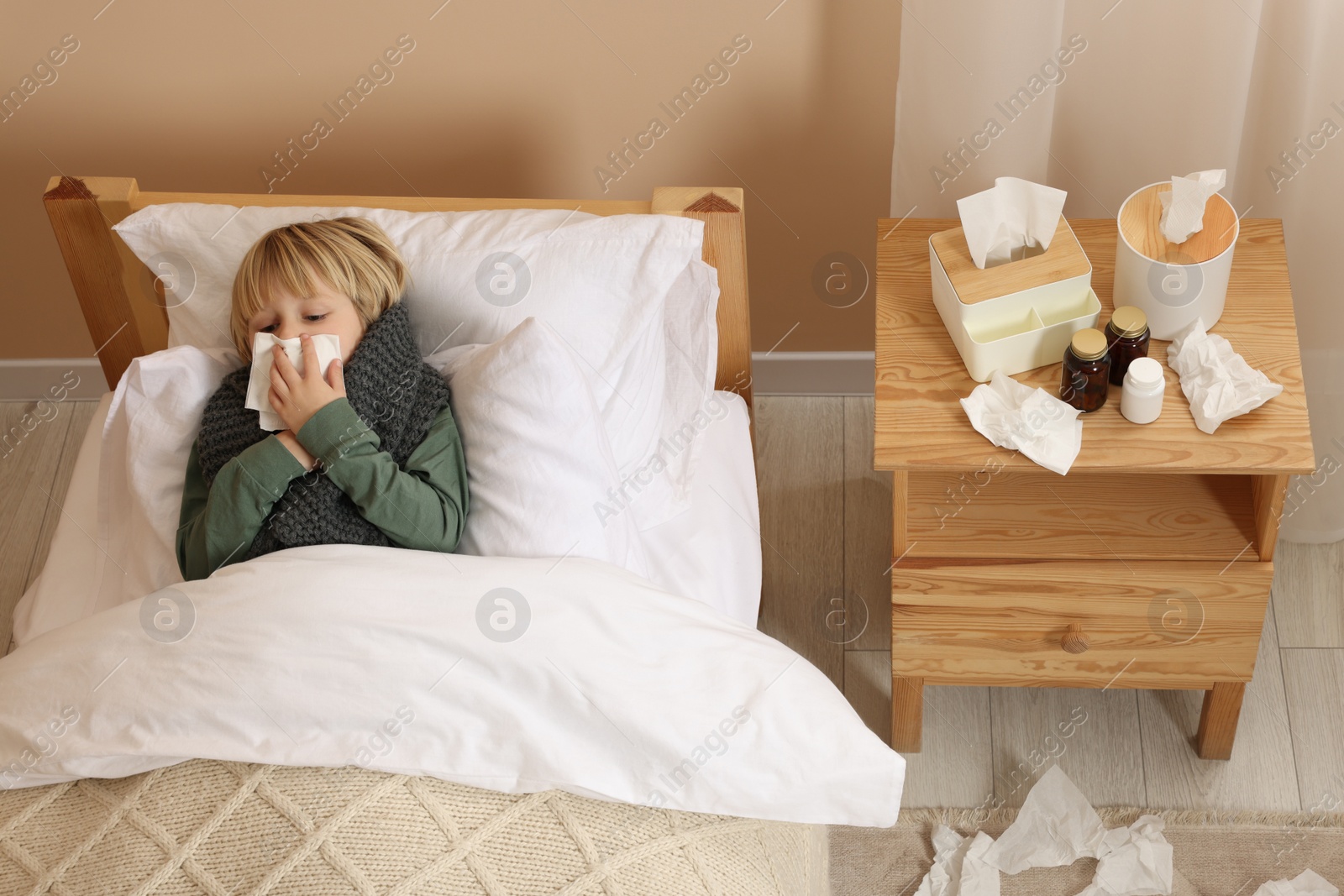 Photo of Boy blowing nose in tissue while lying in bed at home, above view. Cold symptoms