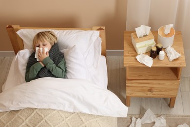 Boy blowing nose in tissue while lying in bed at home, above view. Cold symptoms