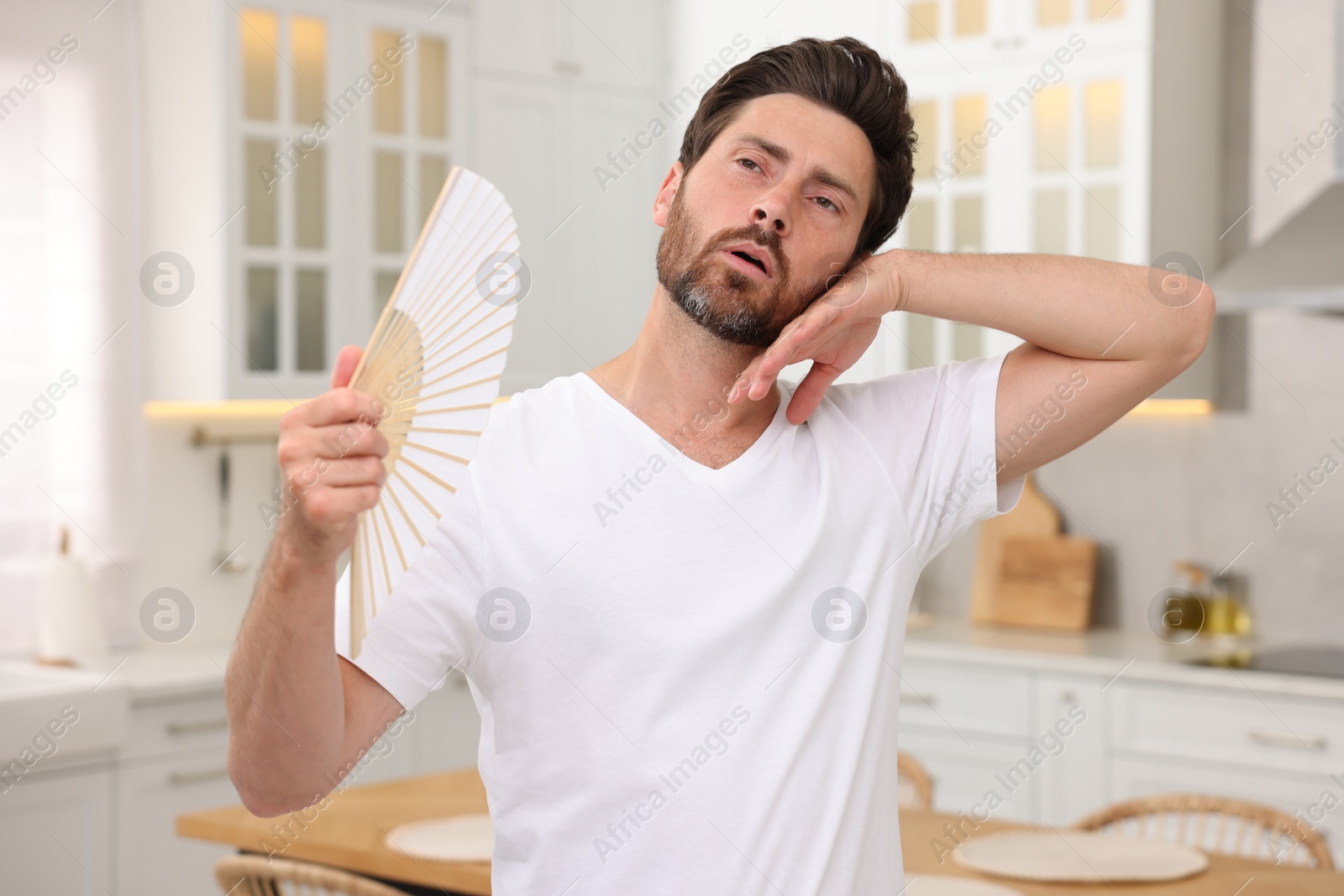 Photo of Bearded man waving white hand fan to cool himself in kitchen