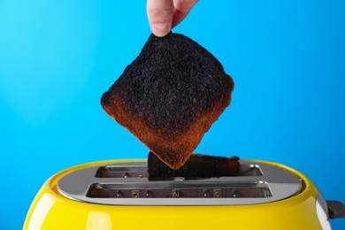 Woman taking off burnt bread from toaster against light blue background, closeup