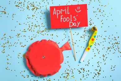 Photo of Sign with phrase April Fools' Day, whoopee cushion and party blower on light blue background, flat lay