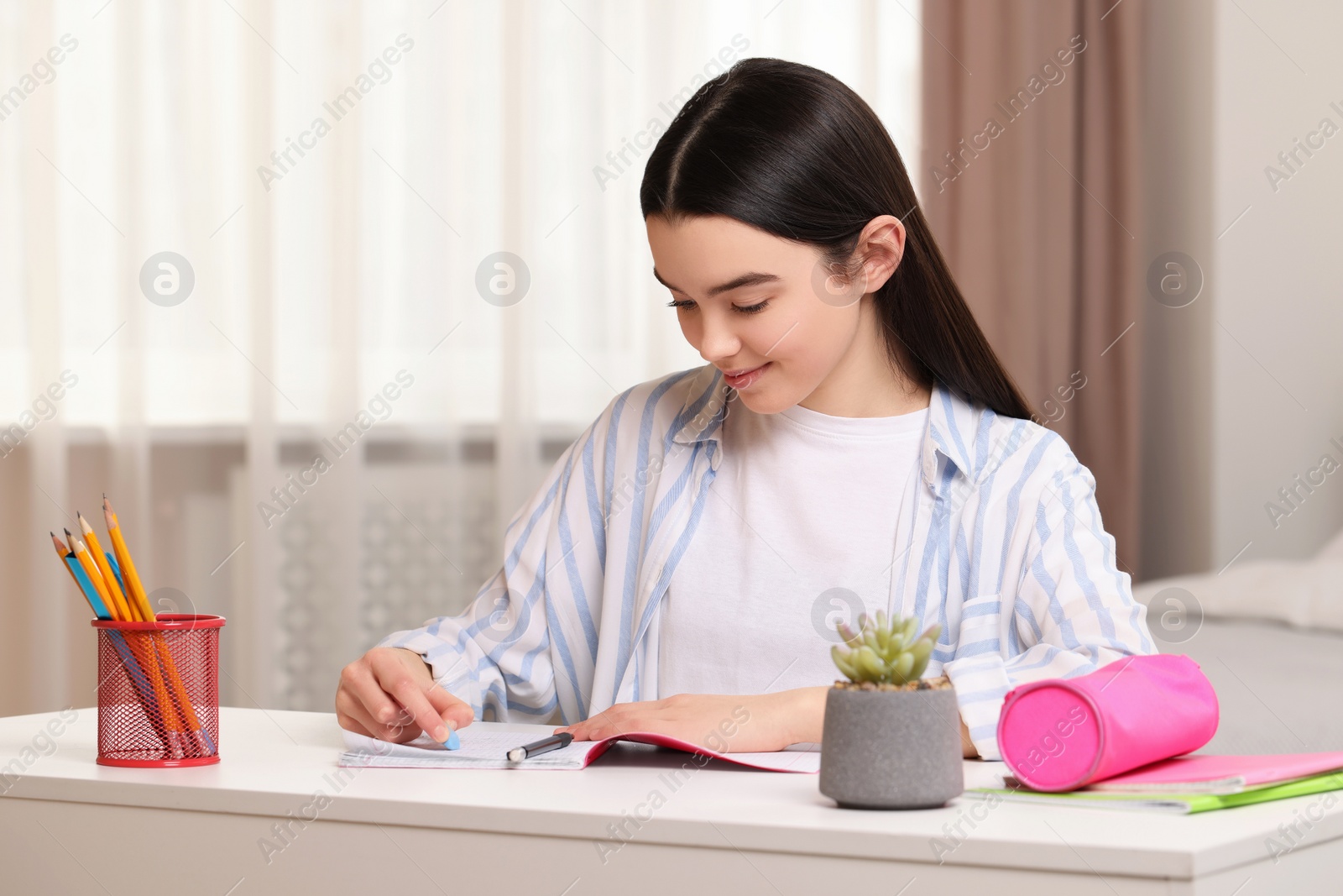 Photo of Teenage girl erasing mistake in her notebook at white desk indoors