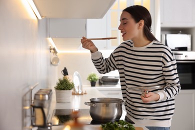Photo of Smiling woman with wooden spoon tasting soup in kitchen