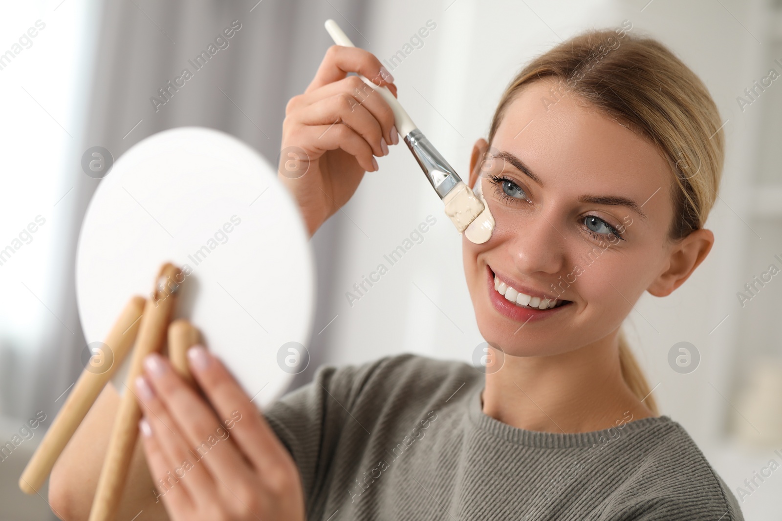 Photo of Young woman applying face mask in front of mirror indoors. Spa treatments