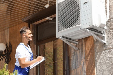 Photo of Professional technician with clipboard near air conditioner outdoors