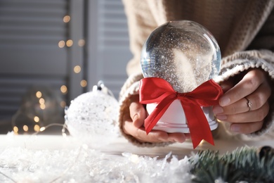 Photo of Woman holding Christmas snow globe with red bow on blurred background, closeup. Space for text