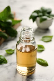 Photo of Glass bottle of basil essential oil and leaves on white  marble table, closeup