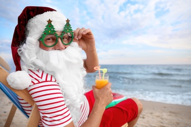 Photo of Santa Claus in party glasses with cocktail relaxing on beach. Christmas vacation