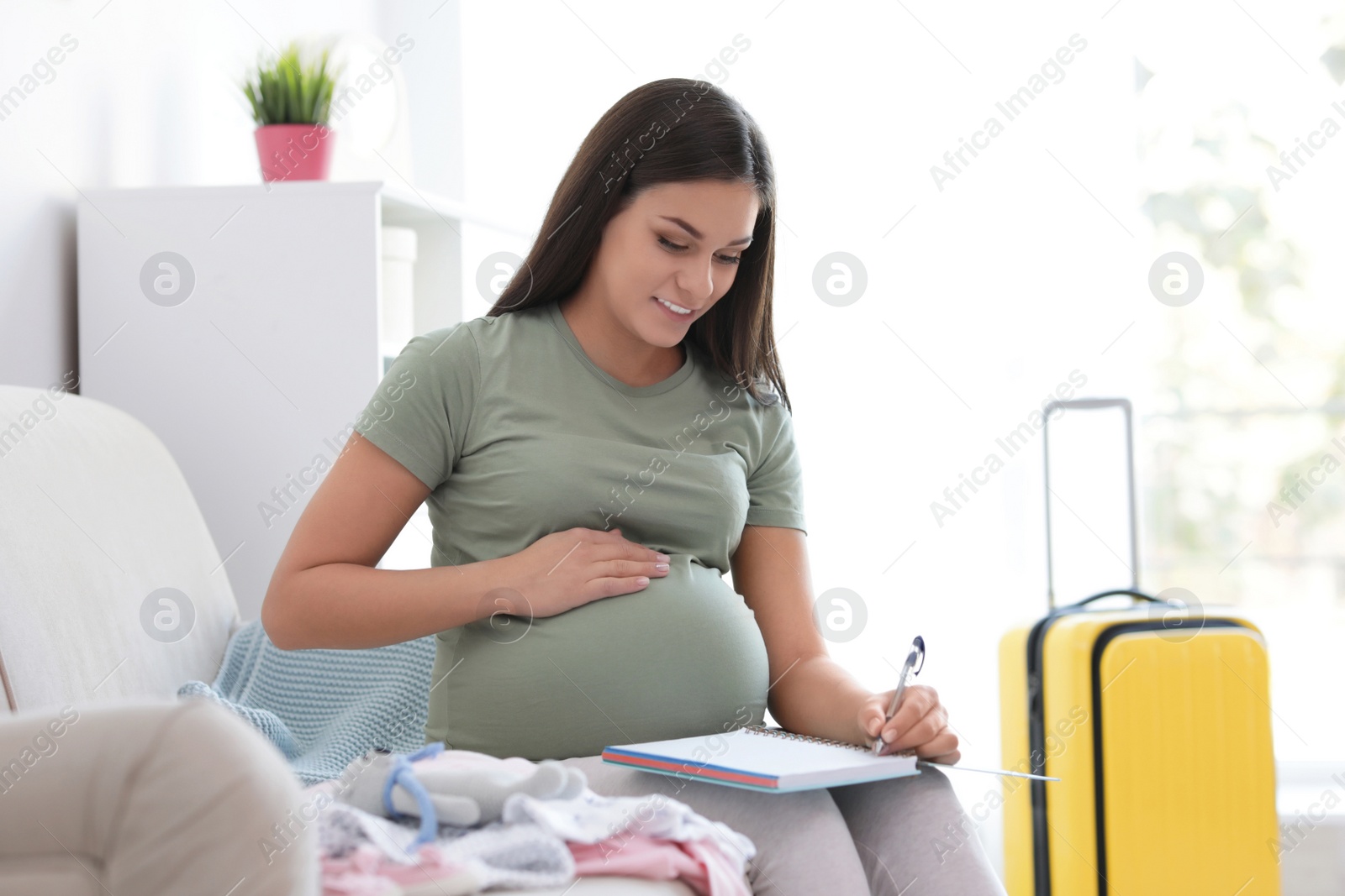 Photo of Pregnant woman making list while packing suitcase for maternity hospital in living room