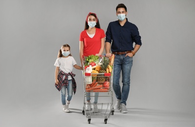 Family in medical masks with shopping cart full of groceries on light grey background