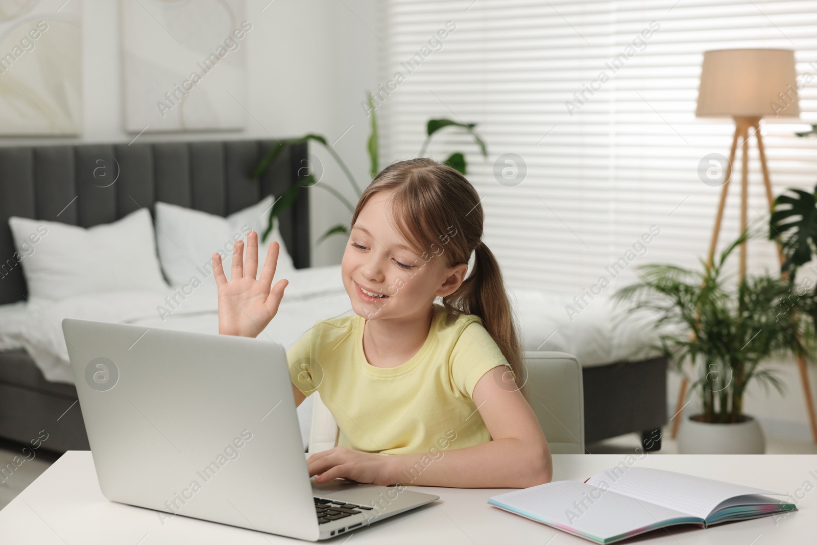 Photo of Cute girl waving hello during online lesson via laptop at white table indoors