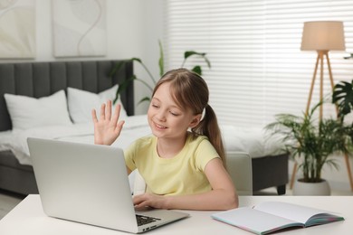 Cute girl waving hello during online lesson via laptop at white table indoors