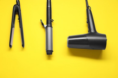 Curling iron, hair straightener and dryer on yellow background, flat lay