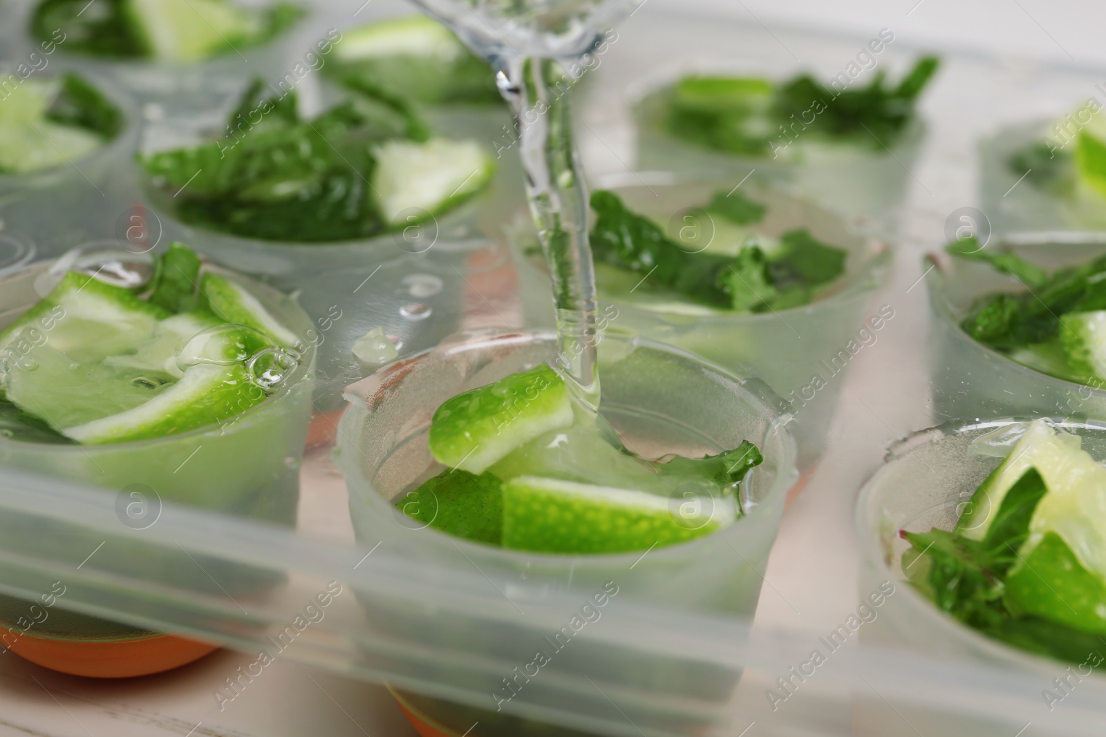 Photo of Pouring water into ice cube tray with lime slices and mint, closeup