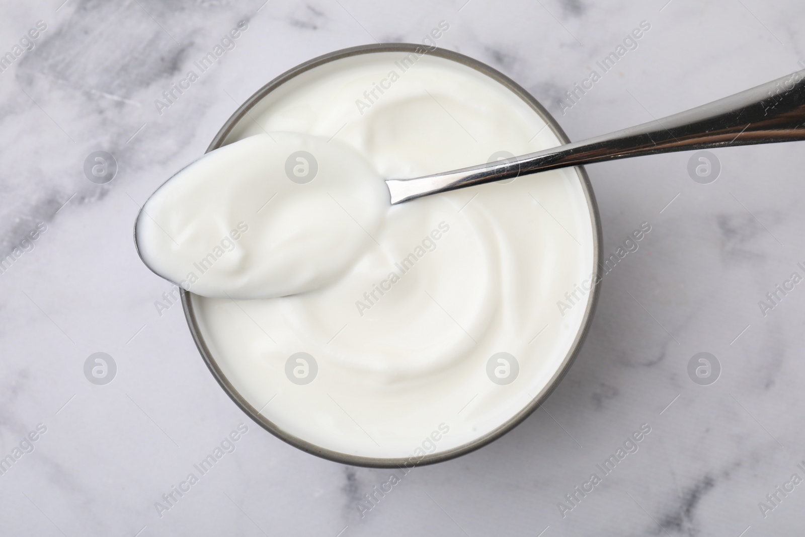 Photo of Delicious natural yogurt in bowl and spoon on white marble table, top view