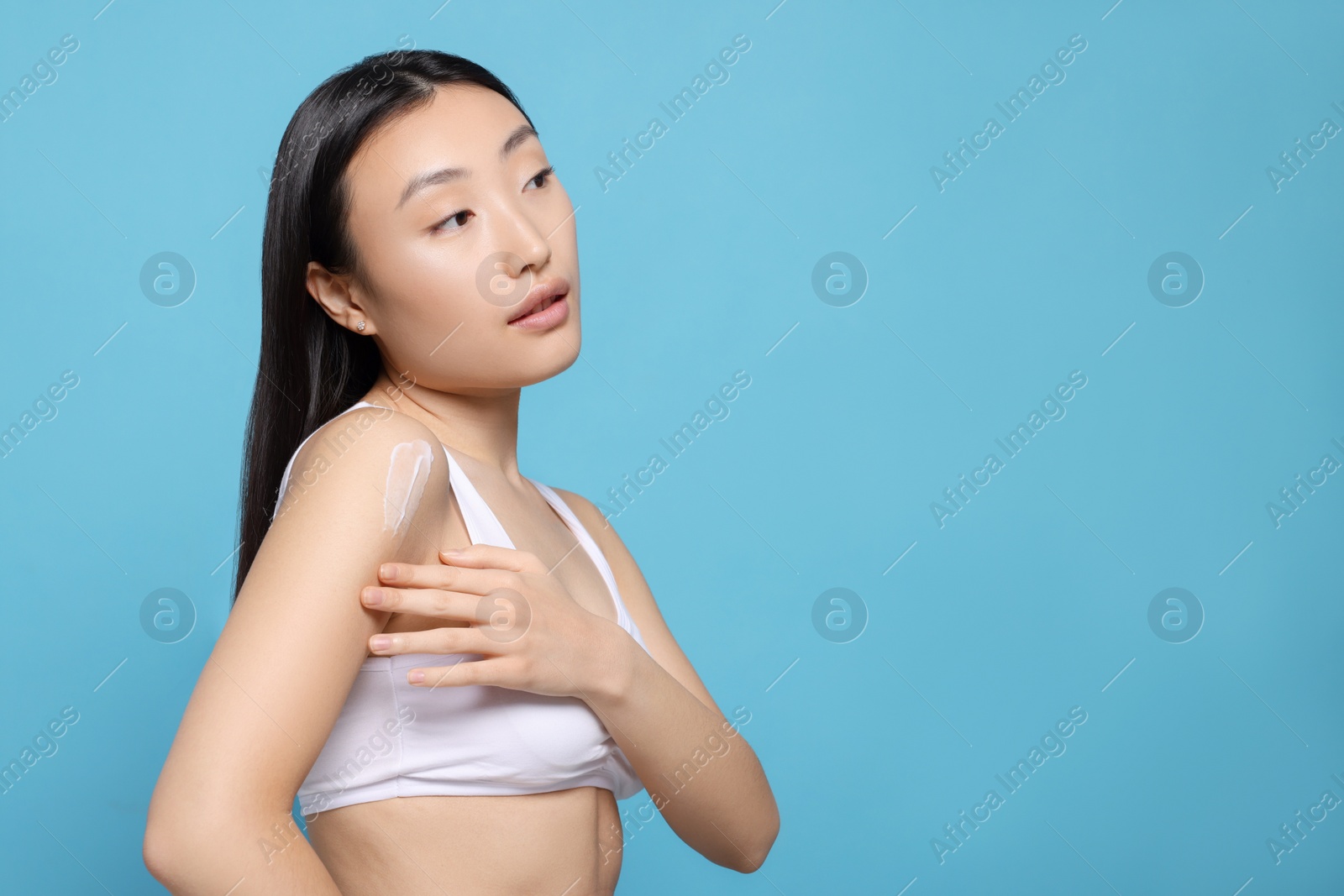 Photo of Beautiful young Asian woman applying body cream onto shoulder on light blue background, space for text