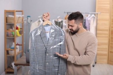 Photo of Dry-cleaning service. Emotional man holding hanger with jacket in plastic bag indoors