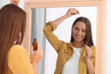 Photo of Beautiful woman applying serum onto hair near mirror indoors