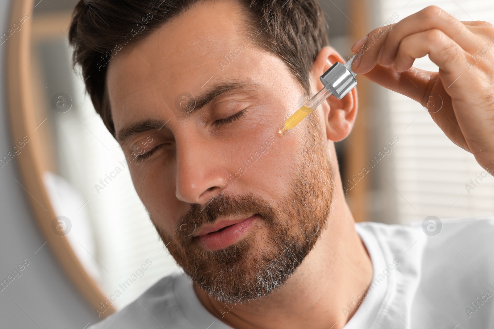 Photo of Handsome man applying cosmetic serum onto his face indoors, closeup