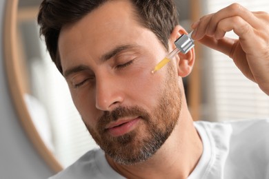 Photo of Handsome man applying cosmetic serum onto his face indoors, closeup