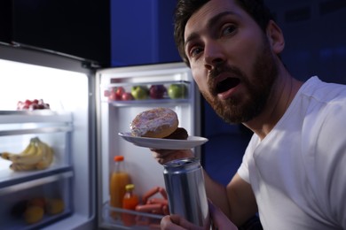 Photo of Man with donuts and drink near refrigerator in kitchen at night, closeup. Bad habit