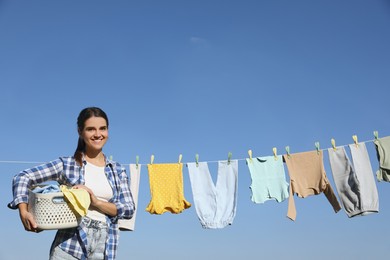 Photo of Smiling woman holding basket with baby clothes near washing line for drying against blue sky outdoors