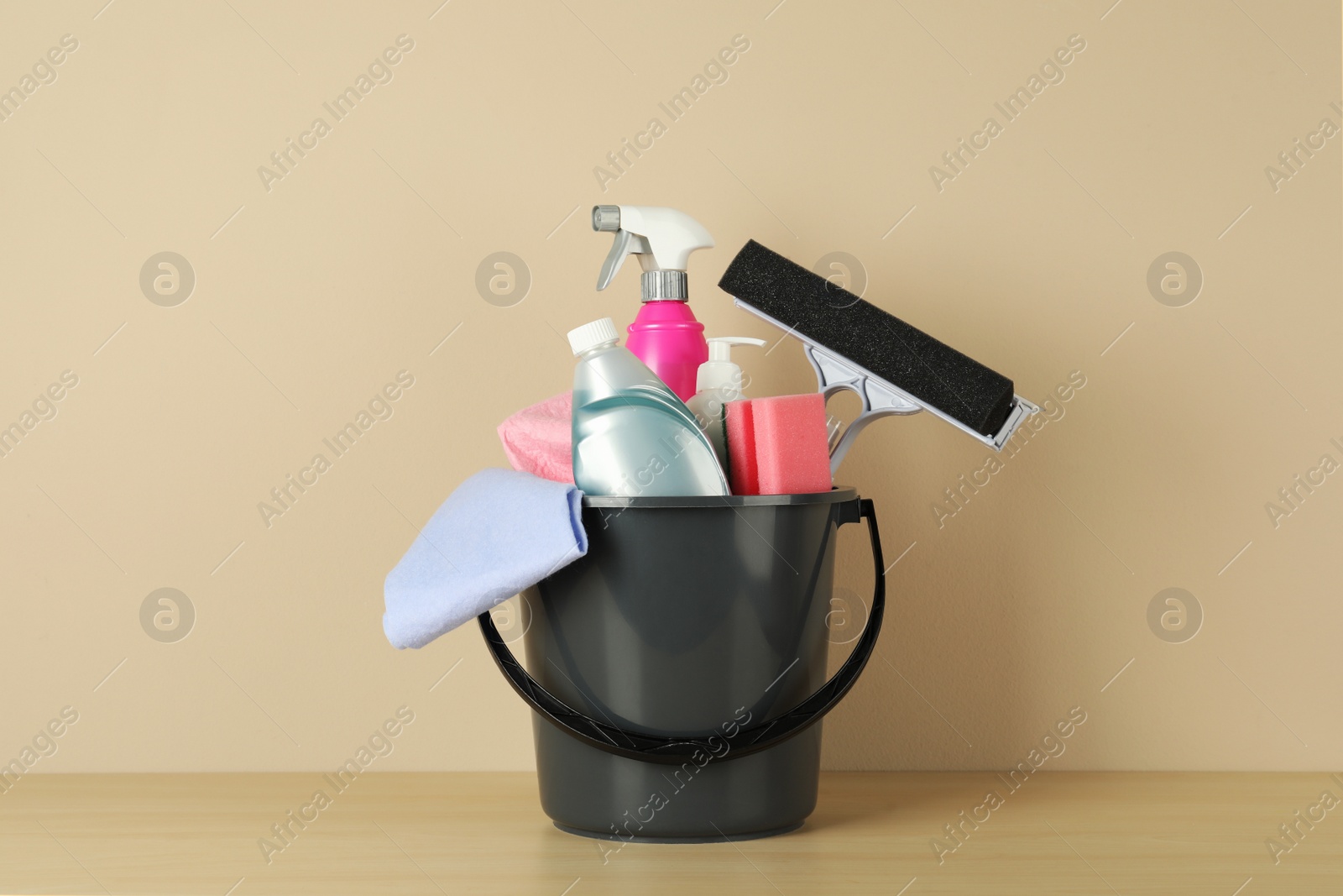 Photo of Bucket with different cleaning supplies on wooden floor near beige wall