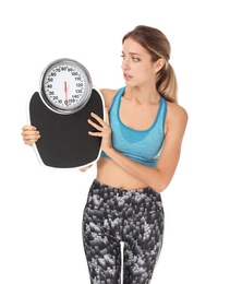 Worried young woman in sportswear holding bathroom scales on white background. Weight loss diet