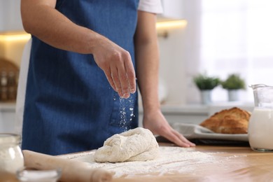 Photo of Making bread. Man sprinkling flour onto dough at wooden table in kitchen, closeup