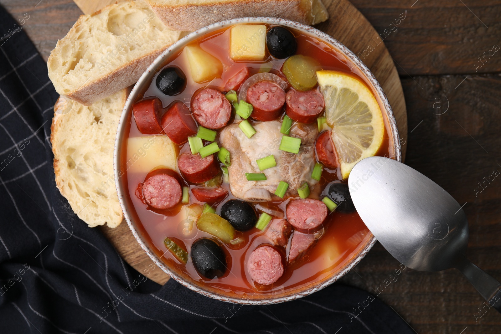 Photo of Meat solyanka soup with thin dry smoked sausages in bowl and bread slices on wooden table, flat lay