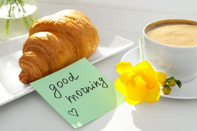 Photo of Cup of aromatic coffee with croissant, beautiful yellow freesia and Good Morning note on windowsill