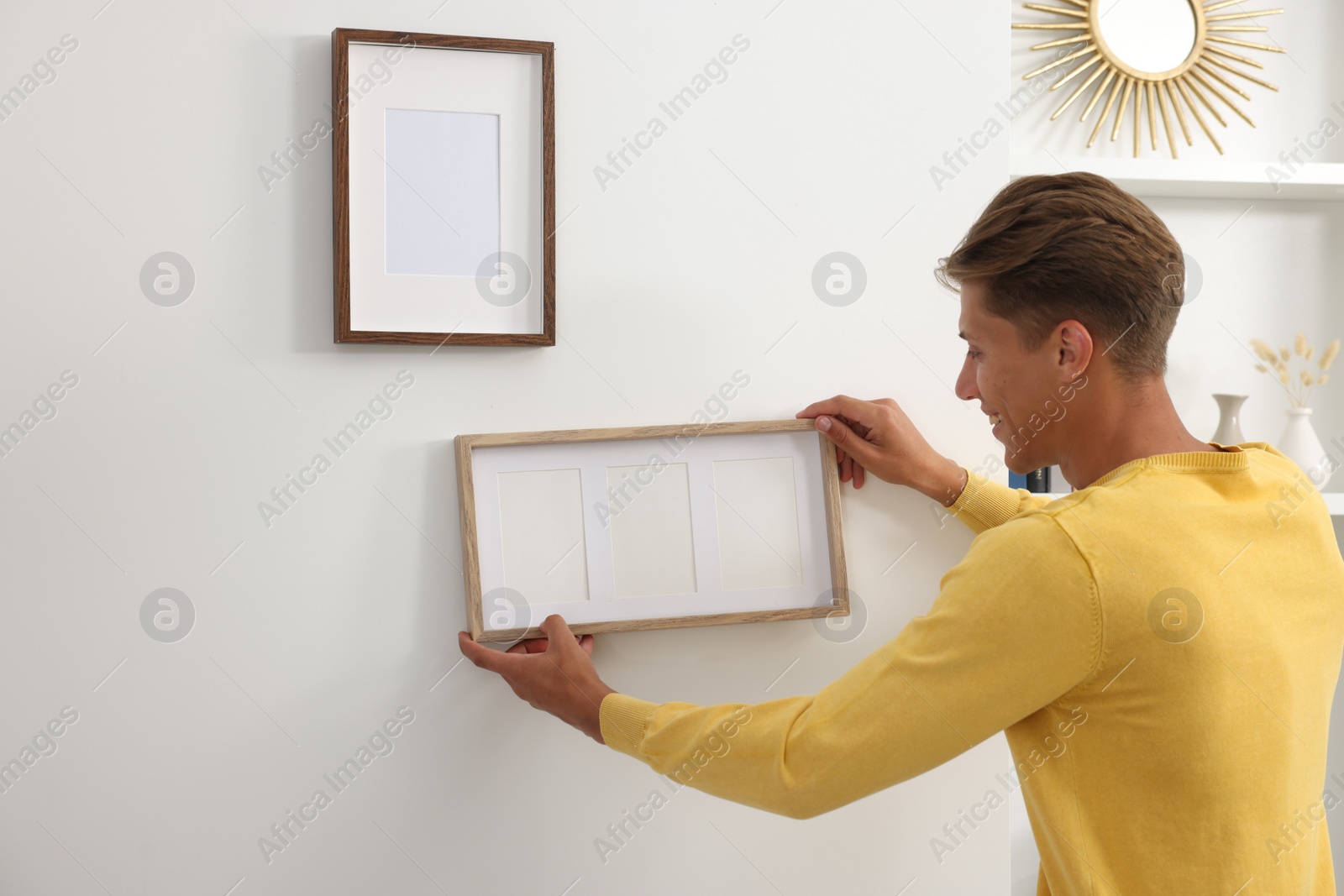 Photo of Young man hanging picture frames on white wall indoors