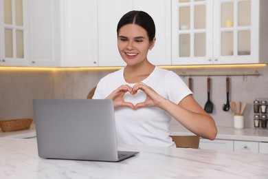Happy young woman having video chat via laptop and making heart at table in kitchen