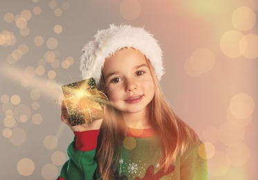Image of Cute child in Santa hat with Christmas gift on color background