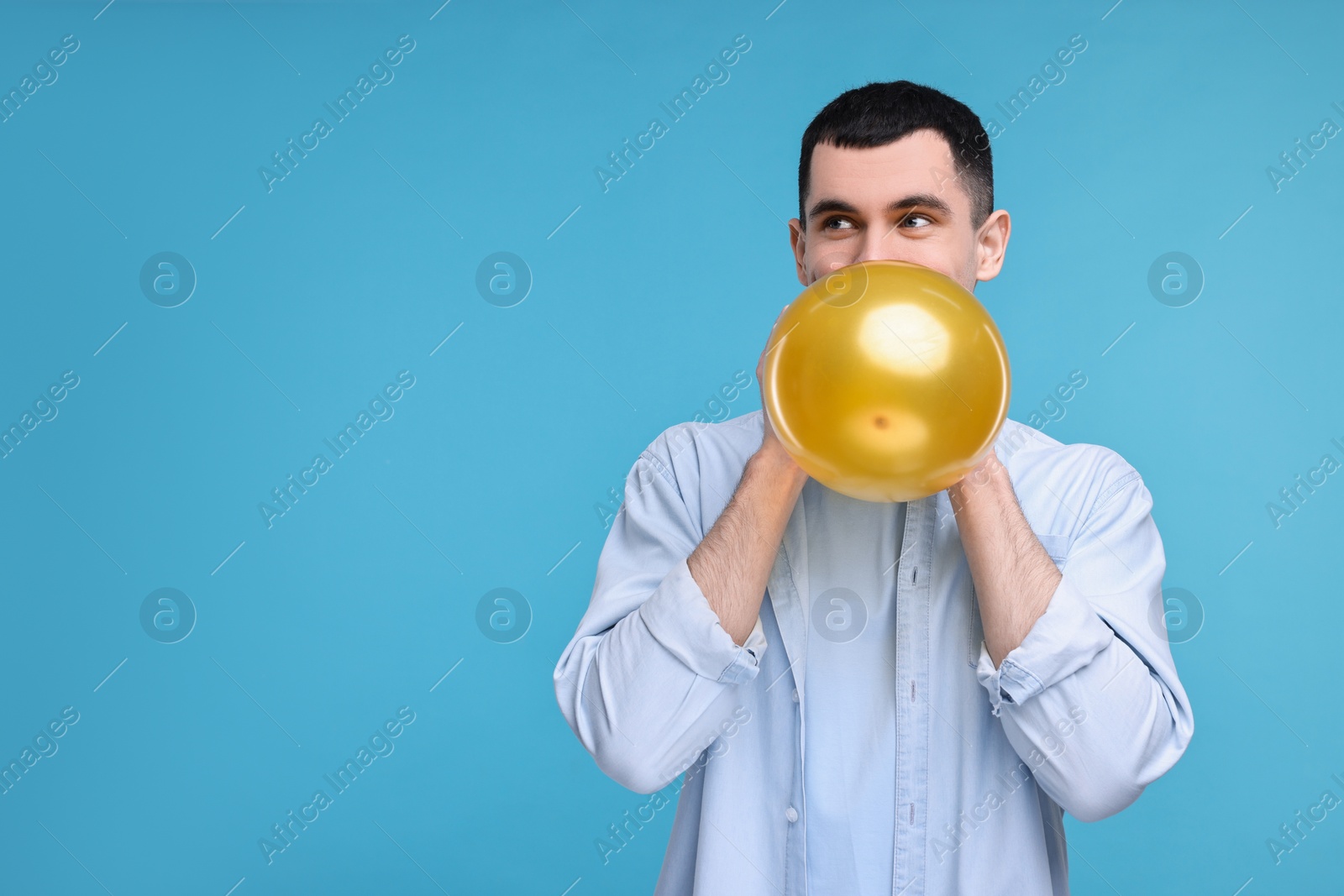 Photo of Young man inflating golden balloon on light blue background. Space for text