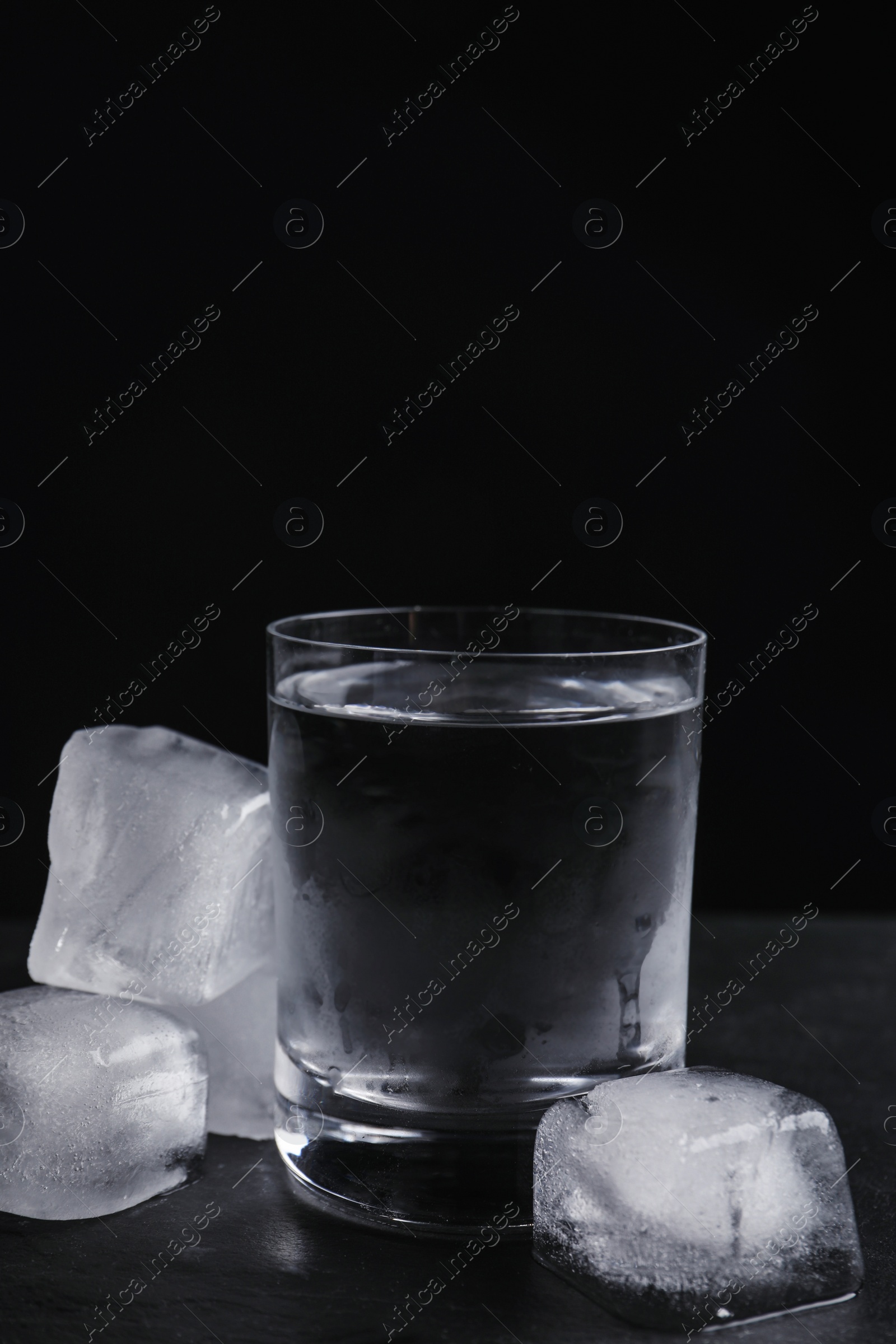 Photo of Vodka in shot glass with ice on table against black background